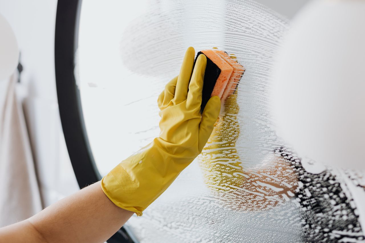 about-us-01 Personne anonyme portant des gants en caoutchouc jaunes et utilisant une simple éponge pour nettoyer un miroir dans une salle de bain
